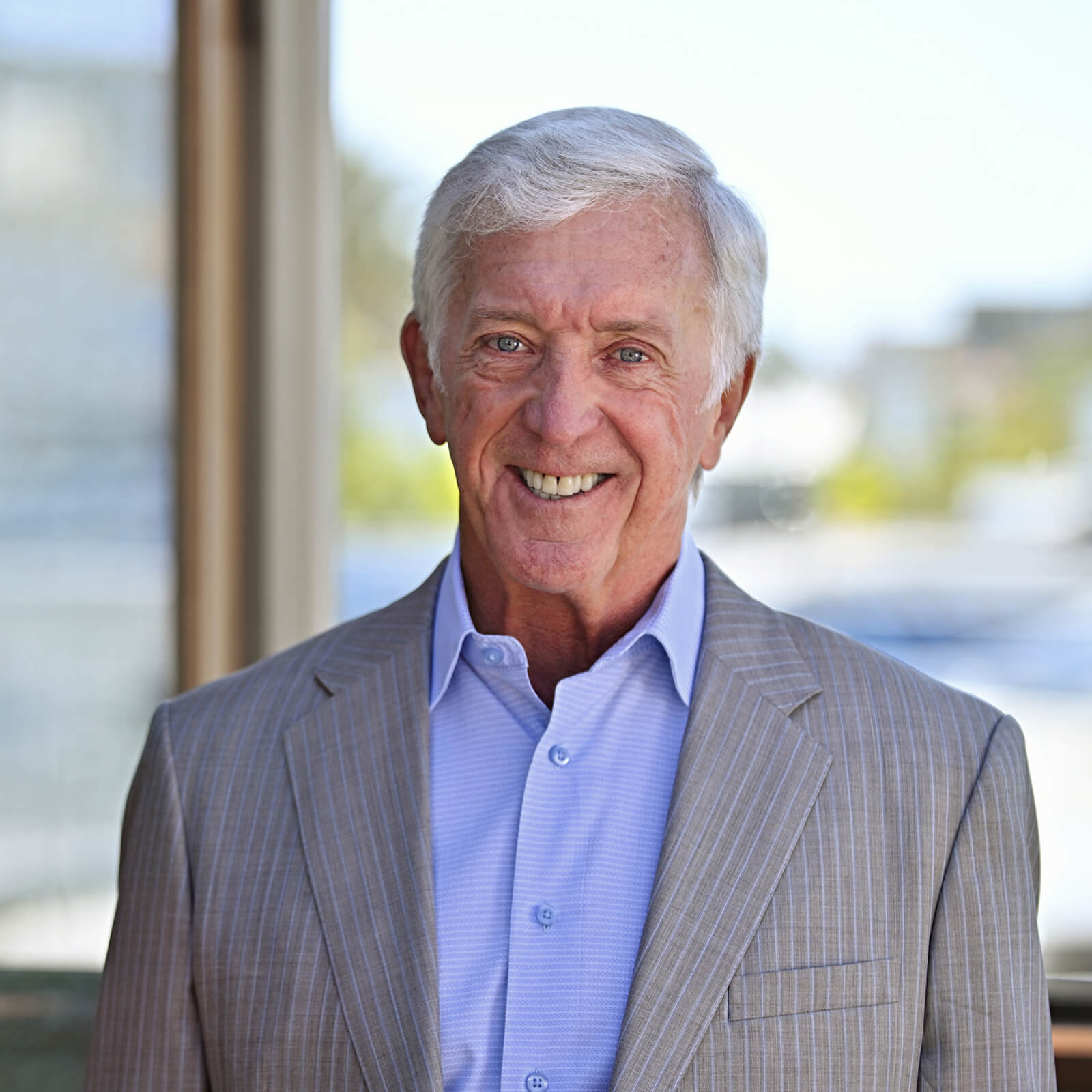 An older man with white hair smiles while wearing a light gray pinstripe suit jacket and a light blue dress shirt. He stands indoors in front of large windows with a blurred outdoor background.