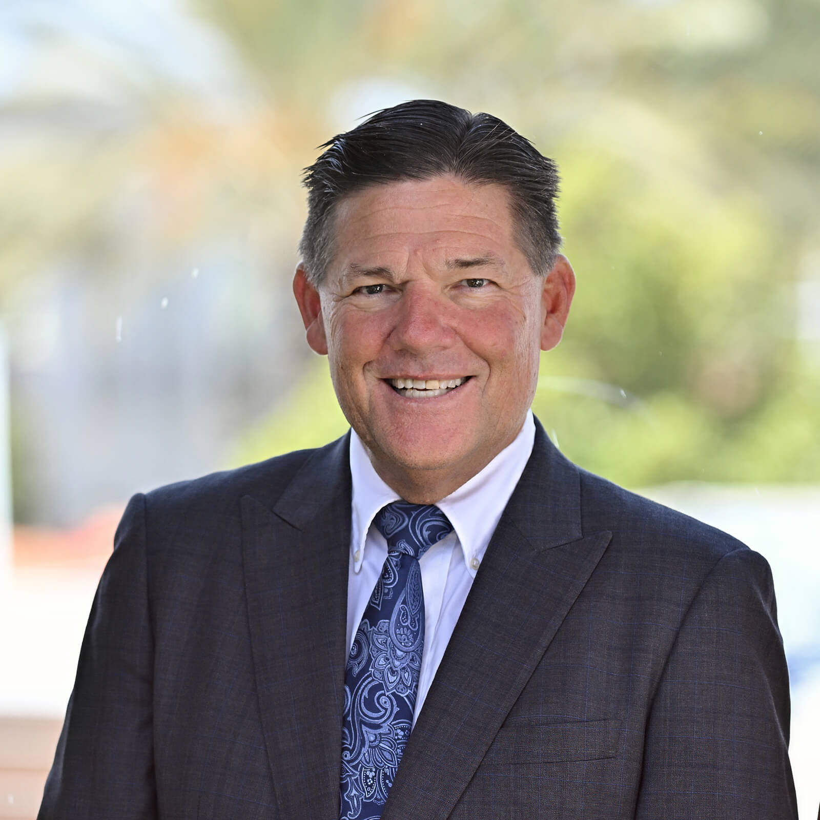 A man in a dark suit, white shirt, and patterned blue tie smiles at the camera against a blurred outdoor background with greenery and soft light.