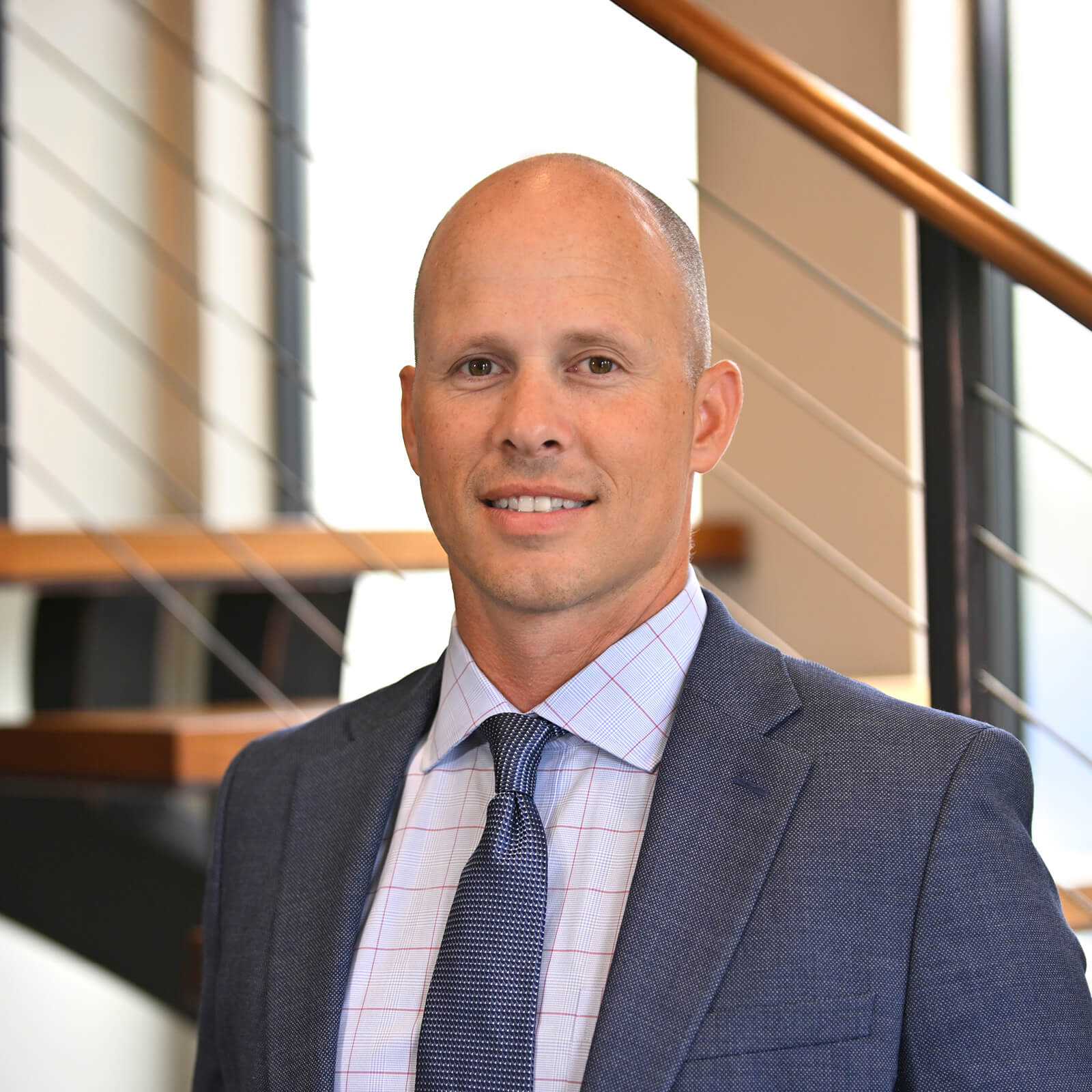 A man in a blue suit and patterned tie stands indoors in front of stairs with metal railings, smiling slightly at the camera. The background is bright with natural light.