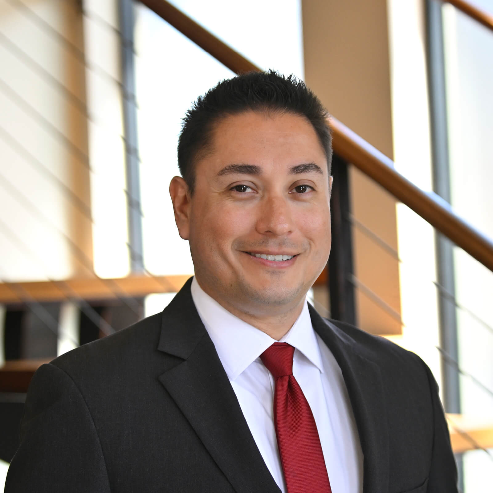 A man in a dark suit, white shirt, and red tie smiles at the camera while standing indoors in front of a staircase with wooden railings and large windows in the background.