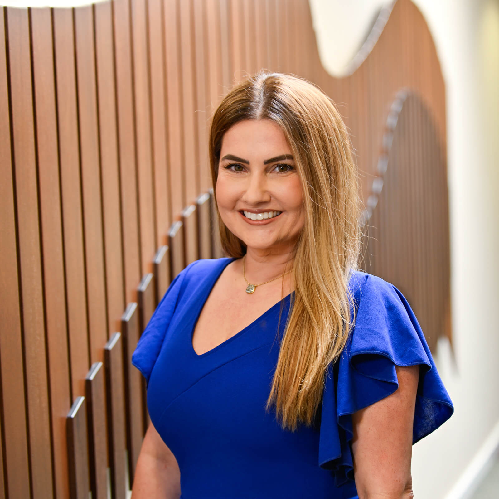 A woman with long, straight brown hair wearing a royal blue dress stands smiling in front of a modern wooden slat wall with a wavy design.