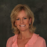 A woman with blonde, shoulder-length hair smiles at the camera. She is wearing a light pink blouse with a ruffled collar and sparkly earrings. The background is dark and softly blurred.