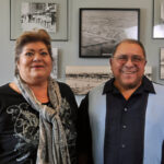 Two adults stand side by side smiling, in front of a wall decorated with black and white photos. The woman wears a patterned scarf and the man wears glasses and a collared shirt.