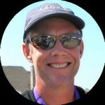 A smiling person wearing a dark baseball cap, sunglasses, and a purple shirt stands outdoors on a sunny day, with part of a building visible in the background. The photo is framed in a circular border.