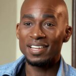 A smiling man with a shaved head and short goatee, wearing a light blue collared shirt, is looking slightly off-camera. The background is softly blurred indoors.