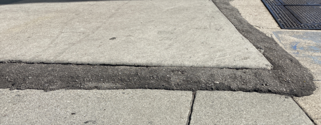 A section of sidewalk shows a raised patch of concrete with uneven, rough asphalt filling the gap between two sidewalk slabs.