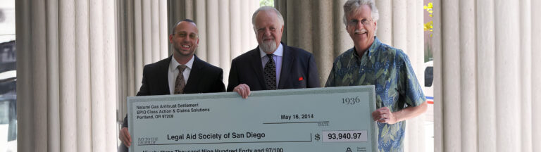 Three men stand outdoors in front of columns, smiling and holding a large ceremonial check made out to the Legal Aid Society of San Diego for $93,949.97, dated May 16, 2014.