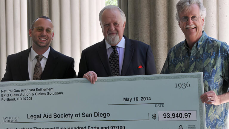 Three men stand outdoors in front of columns, smiling and holding a large ceremonial check made out to the Legal Aid Society of San Diego for $93,949.97, dated May 16, 2014.