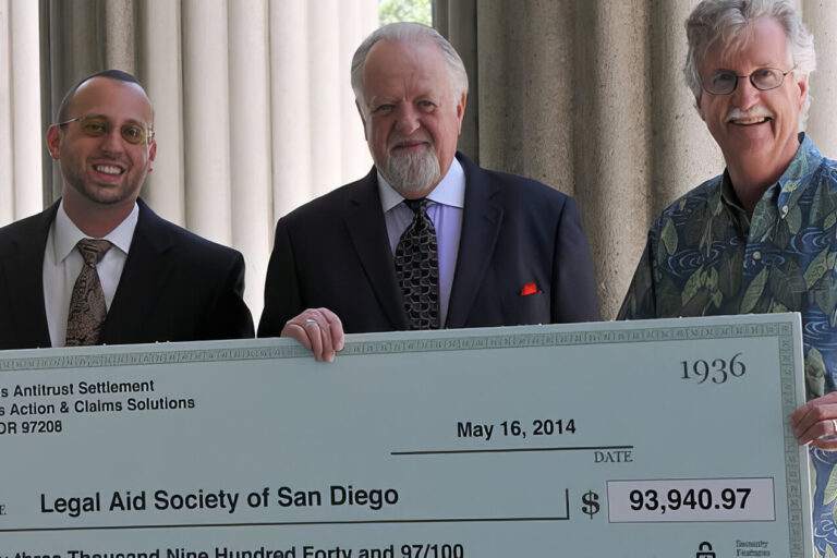 Three men stand outdoors in front of columns, smiling and holding a large ceremonial check made out to the Legal Aid Society of San Diego for $93,949.97, dated May 16, 2014.