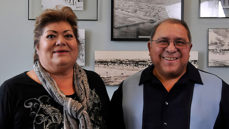 A smiling woman and man stand side by side indoors, in front of a wall decorated with black and white photographs.