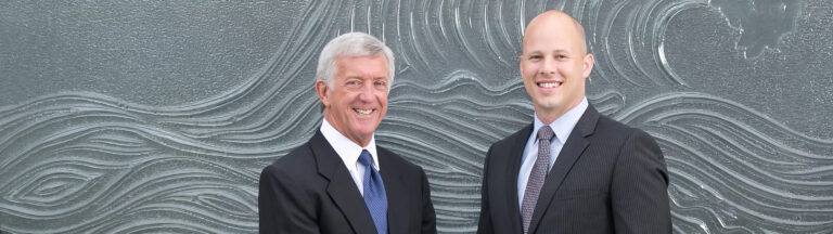 Two men in business suits stand side by side, smiling, in front of a textured wall with wave-like patterns.