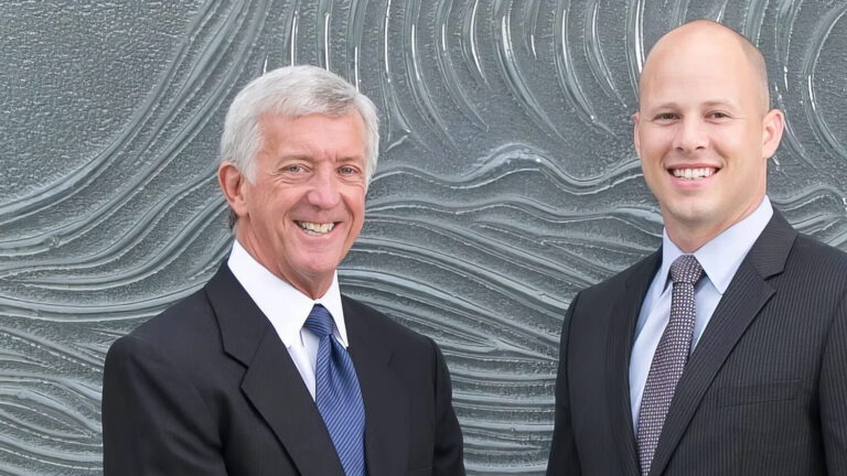 Two men in business suits stand side by side, smiling, in front of a textured wall with wave-like patterns.