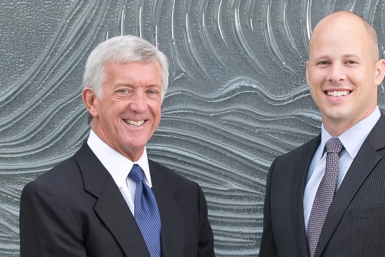 Two men in business suits stand side by side, smiling, in front of a textured wall with wave-like patterns.