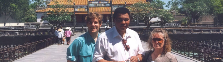 Three people stand together on a bridge in front of a traditional Asian building with a yellow-tiled roof and trees around it. The weather is sunny and clear. Other visitors are seen in the background.