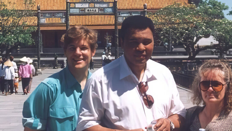 Three people stand together on a bridge in front of a traditional Asian building with a yellow-tiled roof and trees around it. The weather is sunny and clear. Other visitors are seen in the background.
