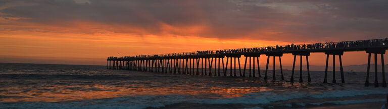 A long pier stretches over the ocean at sunset, with silhouettes of people standing along its length. The sky glows orange and purple, reflecting warm colors onto the waves below.