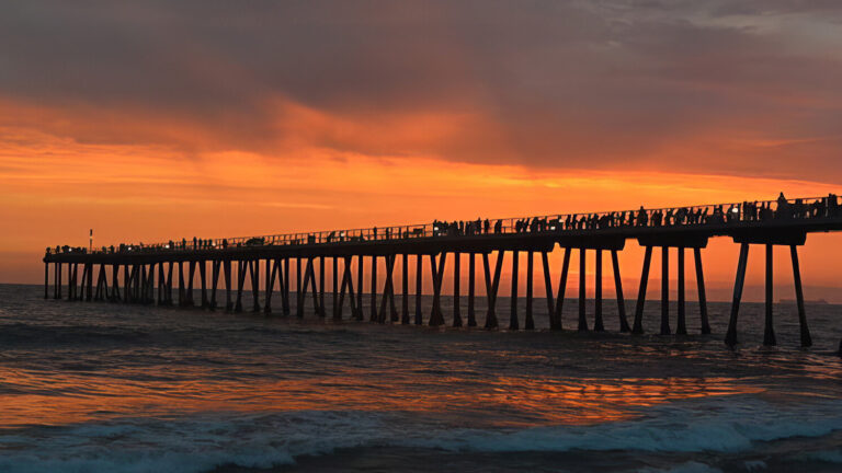 A long pier stretches over the ocean at sunset, with silhouettes of people standing along its length. The sky glows orange and purple, reflecting warm colors onto the waves below.