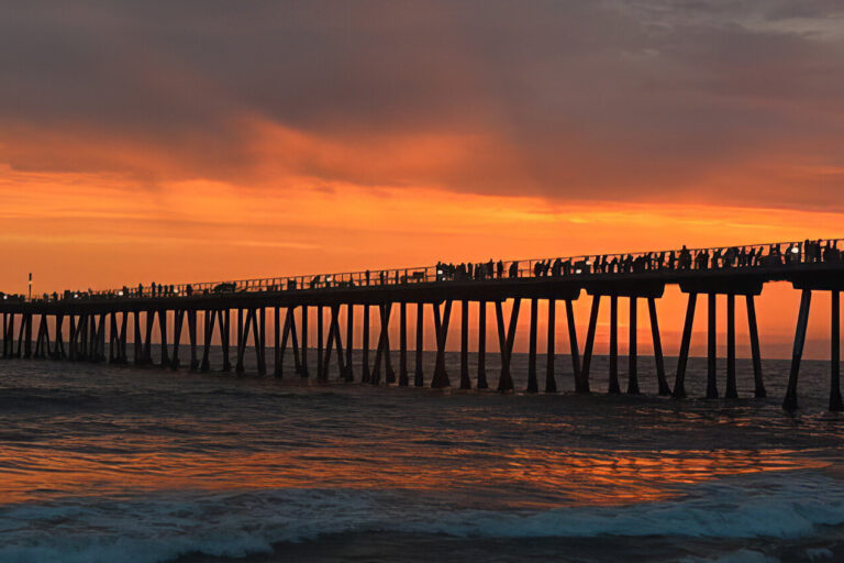 A long pier stretches over the ocean at sunset, with silhouettes of people standing along its length. The sky glows orange and purple, reflecting warm colors onto the waves below.
