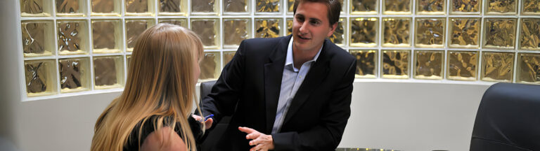A man in a dark suit sits at a desk, talking and gesturing with a pen to a woman with long blonde hair, in an office with glass block walls.