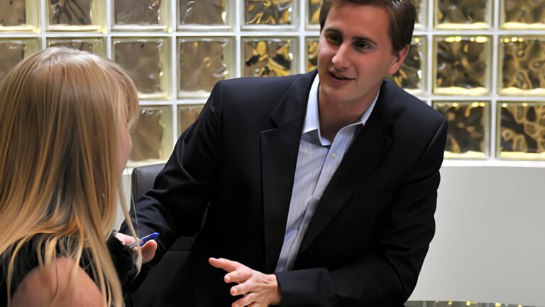 A man in a dark suit sits at a desk, talking and gesturing with a pen to a woman with long blonde hair, in an office with glass block walls.