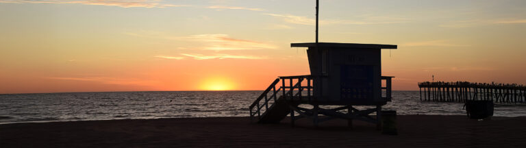 A lifeguard tower is silhouetted against a colorful sunset sky on a sandy beach, with calm ocean waves and a distant pier visible on the right.