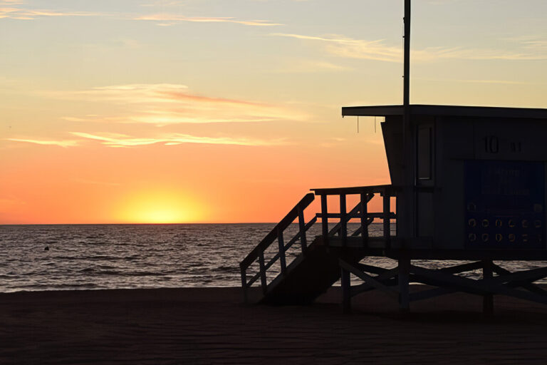 A lifeguard tower is silhouetted against a colorful sunset sky on a sandy beach, with calm ocean waves and a distant pier visible on the right.