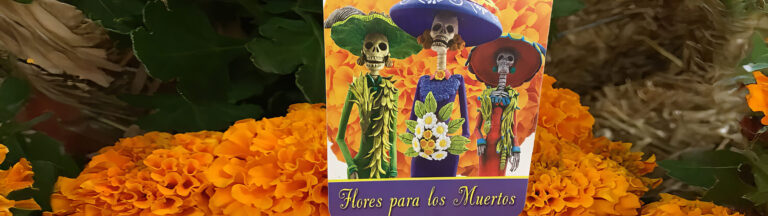 A card with three skeleton figures in colorful clothing and hats stands among orange marigold flowers. The card reads “Flores para los Muertos.”.