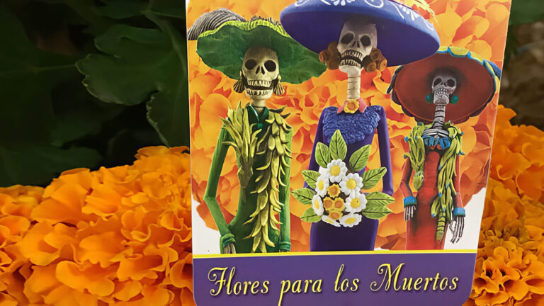 A card with three skeleton figures in colorful clothing and hats stands among orange marigold flowers. The card reads “Flores para los Muertos.”.