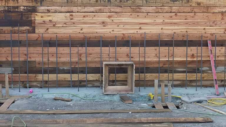 A construction site with a wooden wall, metal rebar grid, open wooden frames, and scattered construction materials, including hoses, planks, and tools on a concrete floor.