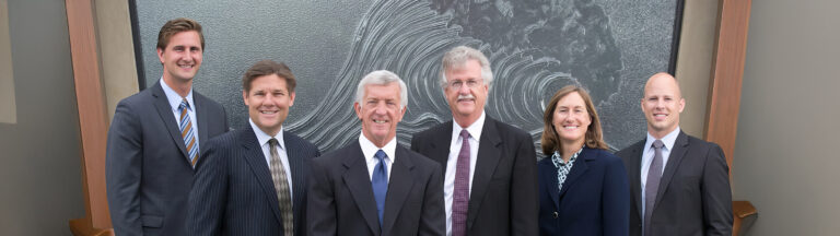Six people in business attire stand side by side, smiling, in front of a large, abstract artwork featuring swirling patterns. Four men in suits and ties and one woman in a suit are visible in the group.