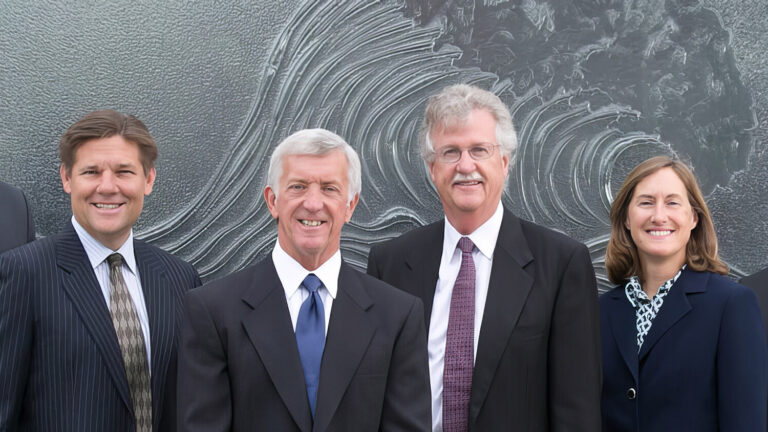 Six people in business attire stand side by side, smiling, in front of a large, abstract artwork featuring swirling patterns. Four men in suits and ties and one woman in a suit are visible in the group.