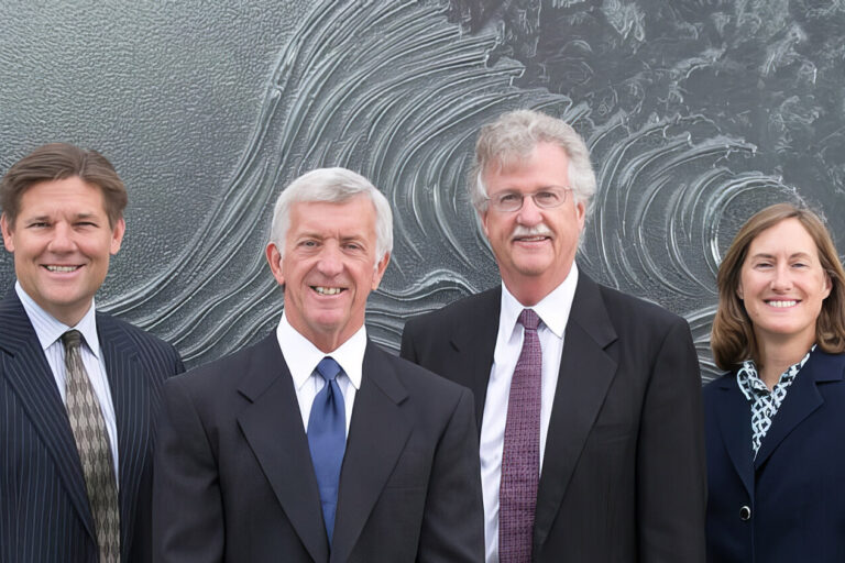 Six people in business attire stand side by side, smiling, in front of a large, abstract artwork featuring swirling patterns. Four men in suits and ties and one woman in a suit are visible in the group.