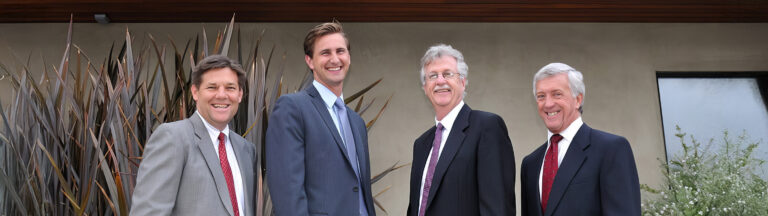 Four men in suits stand outdoors in front of a building, smiling at the camera. The background features tall plants and a window.
