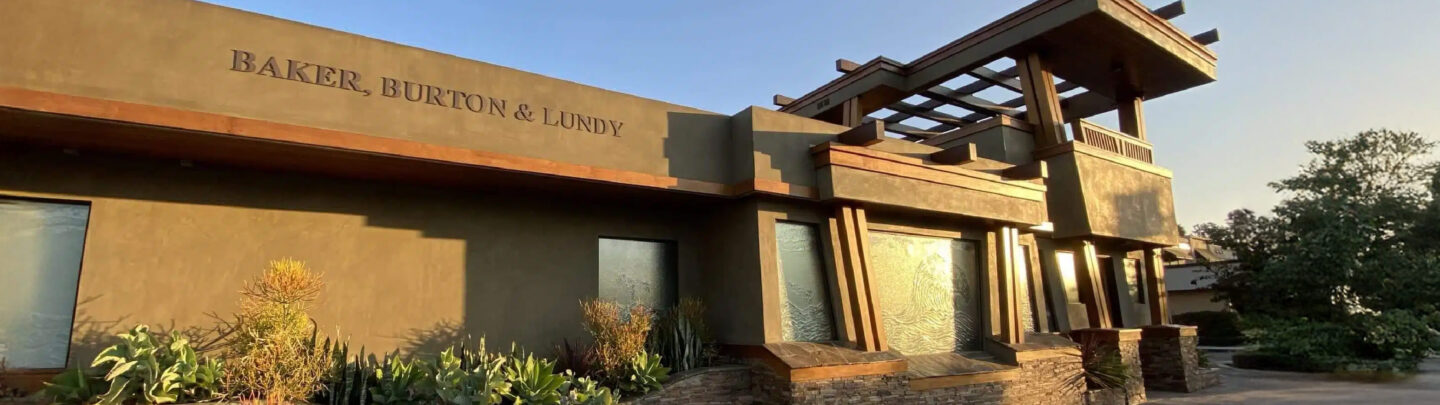 Modern office building with Baker, Burton & Lundy on the facade, large windows, stone accents, and landscaped greenery in the foreground, lit by warm sunlight.