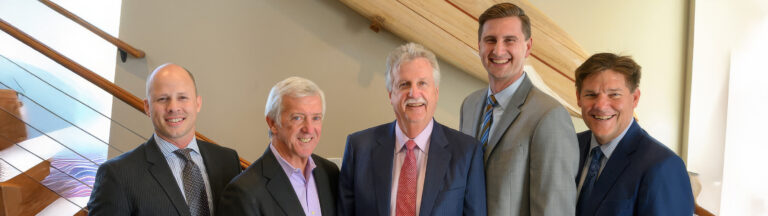 Five men in business suits stand side by side, smiling, indoors in front of a wooden staircase and light-colored wall.
