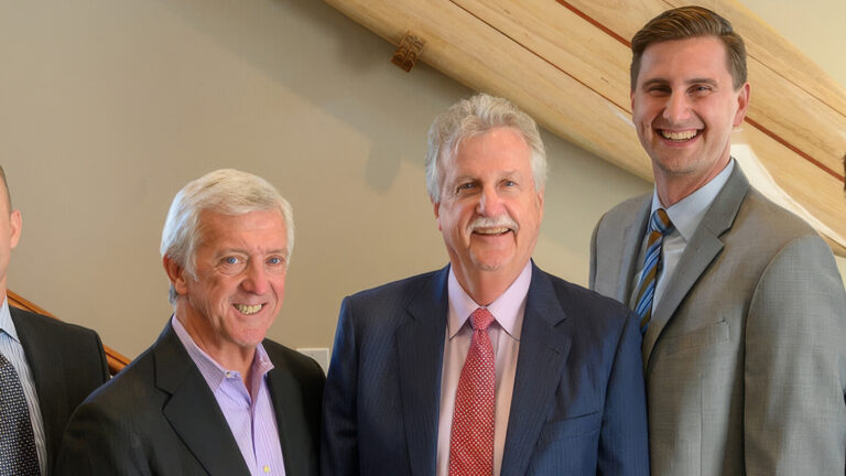 Five men in business suits stand side by side, smiling, indoors in front of a wooden staircase and light-colored wall.