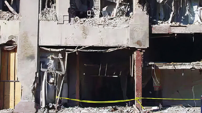 A heavily damaged building with debris scattered around, large holes in the facade, exposed interiors, and yellow caution tape blocking the entrance. The structure appears partially collapsed.