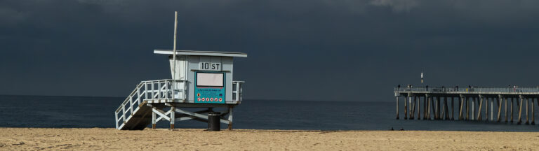 A lifeguard tower labeled 107 stands vacant on a sandy beach, with a dark sky and calm ocean in the background. A pier extends into the water on the right side of the image.