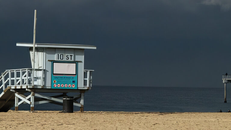 A lifeguard tower labeled 107 stands vacant on a sandy beach, with a dark sky and calm ocean in the background. A pier extends into the water on the right side of the image.