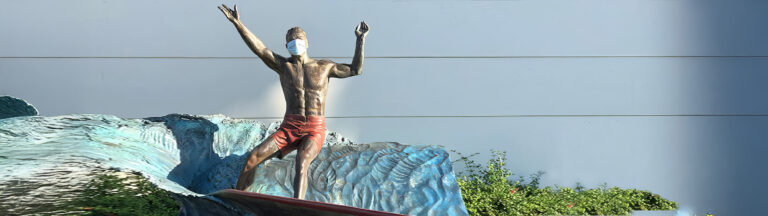 A bronze statue of a surfer wearing red shorts and a face mask stands on a wave in front of a blue-gray wall, with green shrubs at the base.