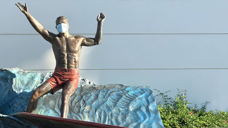 A bronze statue of a surfer wearing red shorts and a face mask stands on a wave in front of a blue-gray wall, with green shrubs at the base.
