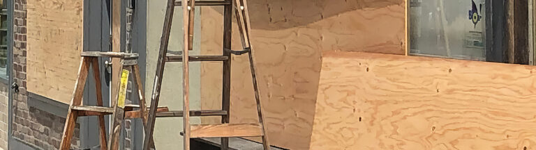 Two wooden ladders stand in front of a building with its windows and door covered by large sheets of plywood. The scene suggests construction or preparation for possible damage.