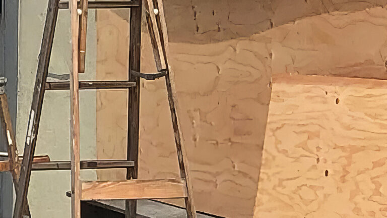 Two wooden ladders stand in front of a building with its windows and door covered by large sheets of plywood. The scene suggests construction or preparation for possible damage.