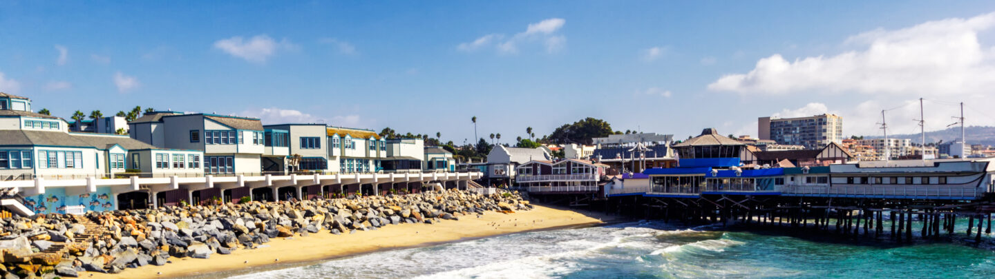 A sunny beach scene with turquoise waves, sandy shore, and a row of buildings on a rocky seawall. A wooden pier extends over the water; city and palm trees are visible in the background under a blue sky.