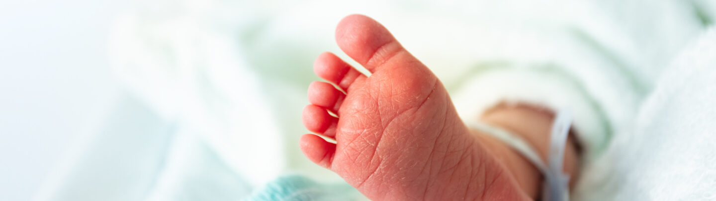 Close-up of a newborn babys foot resting on a soft white blanket, with a hospital identification band around the ankle. The baby is partially covered by the blanket, creating a warm and gentle atmosphere.