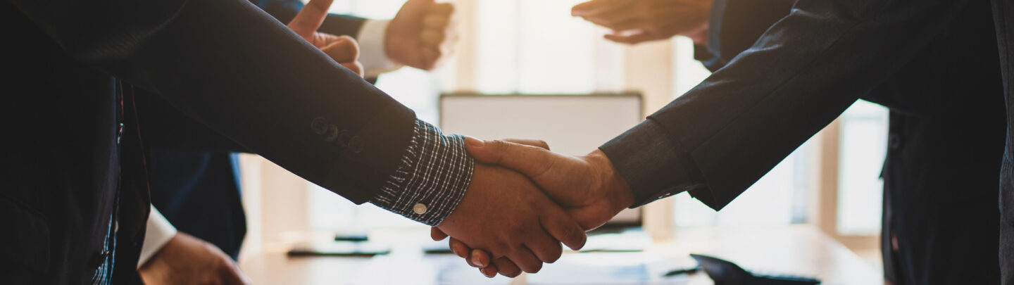 Two people in business attire shake hands across a table with documents and notebooks, while others gesture positively in the background, suggesting a successful meeting or agreement. Sunlight streams in through large windows.