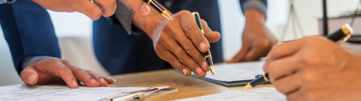 Three people in business attire are gathered around a desk, holding pens and reviewing documents. Only their hands and arms are visible as they gesture and point at papers, suggesting collaboration or discussion.