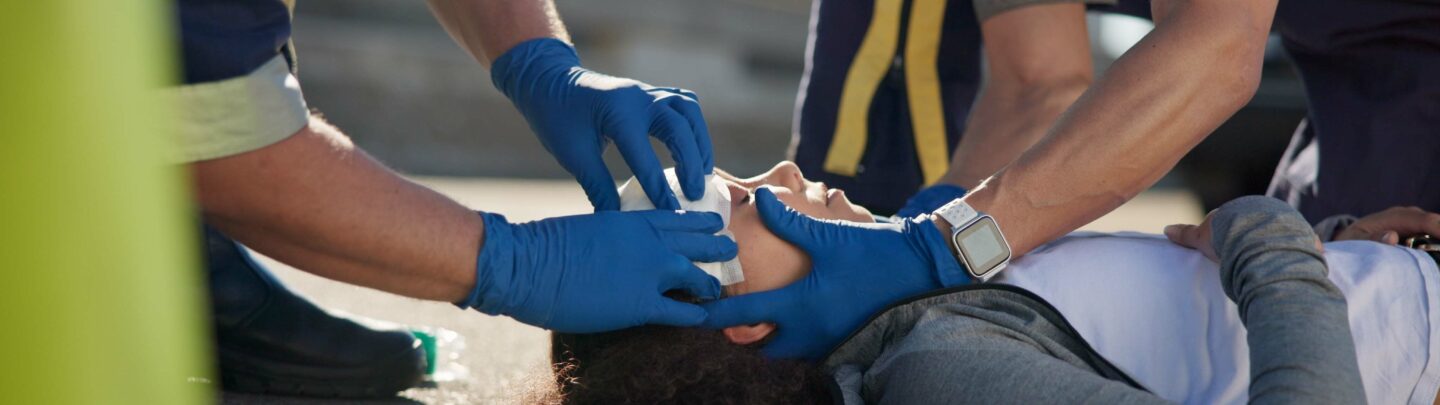 Two emergency responders wearing blue gloves provide medical assistance to a person lying on the ground, stabilizing their head and checking their airway. The scene appears to be outdoors on a paved surface.