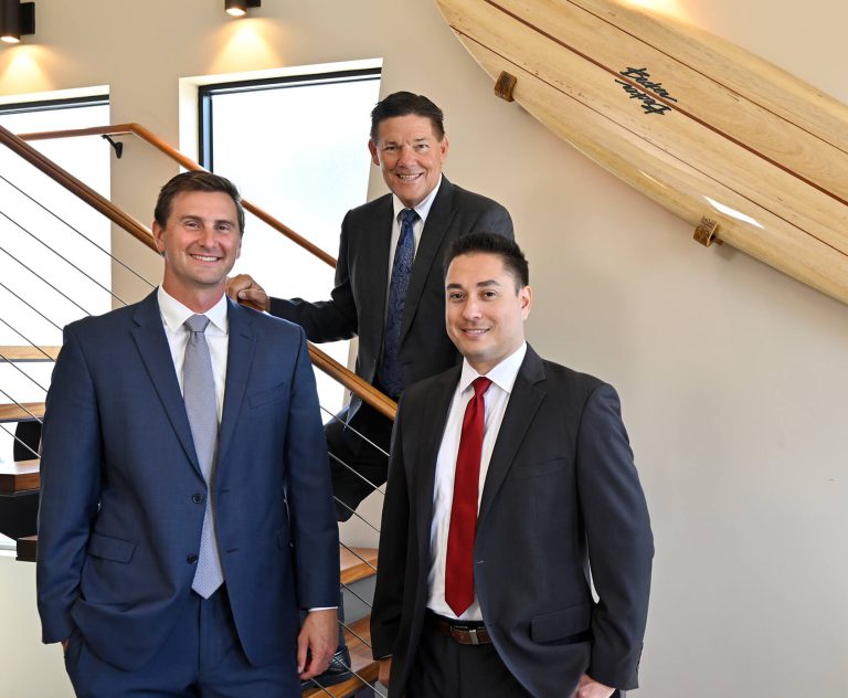 Three men in business suits stand and smile indoors by a staircase and a wooden surfboard mounted on the wall. The setting appears professional and modern with natural light coming through windows.