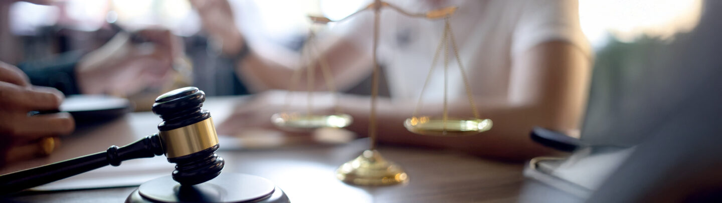 Close-up of a gavel and golden scales of justice on a desk, with blurred people in the background, suggesting a legal or courtroom setting.